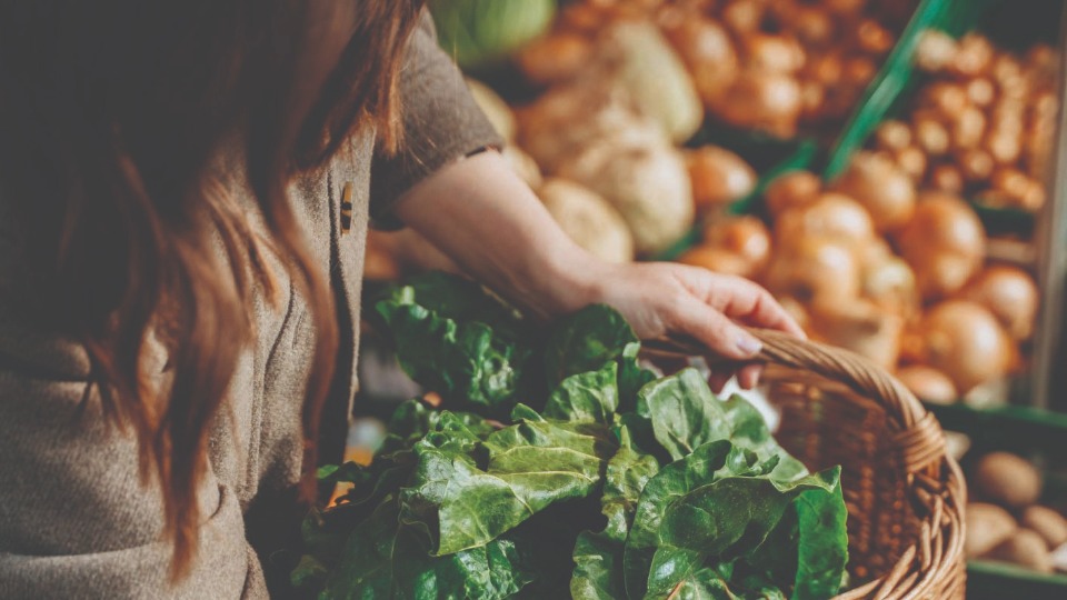 Woman holding basket of lettuce next to more produce.