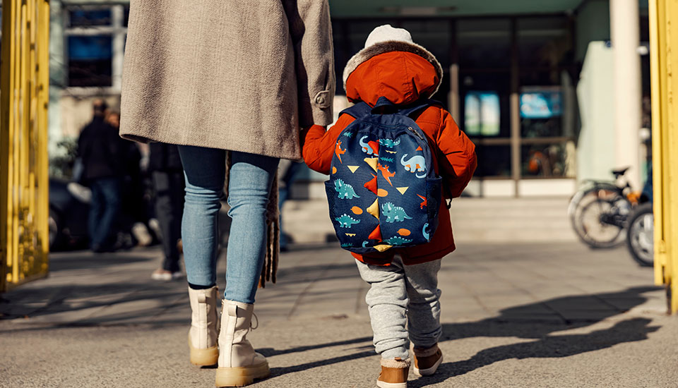 Adult holding young child's hand as they walk into the front door of a school building. The child is wearing a small backpack.