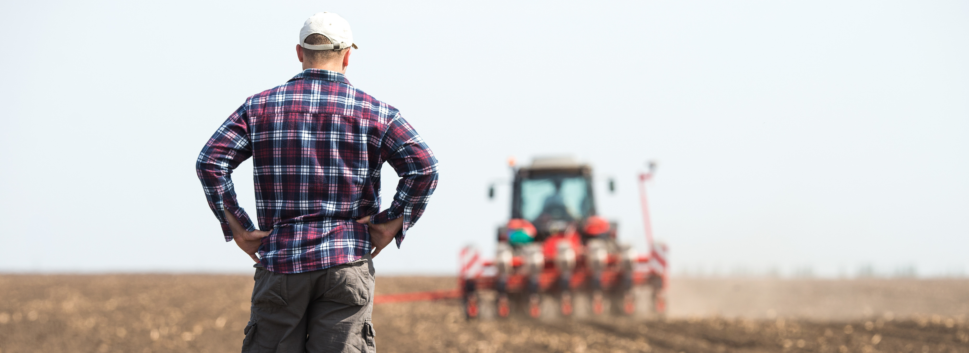 Farmer in plaid shirt looking at tiller in a field, back turned to the camera