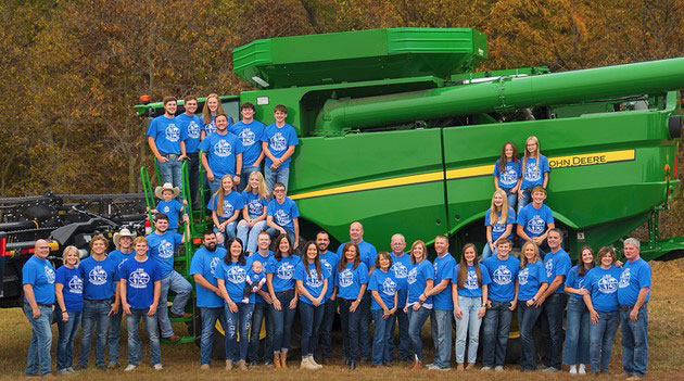 Group photo, numerous members of family wearing blue shirts and standing in front of green John Deere combine Group photo, numerous members of family wearing blue shirts and standing in front of green John Deere combine
