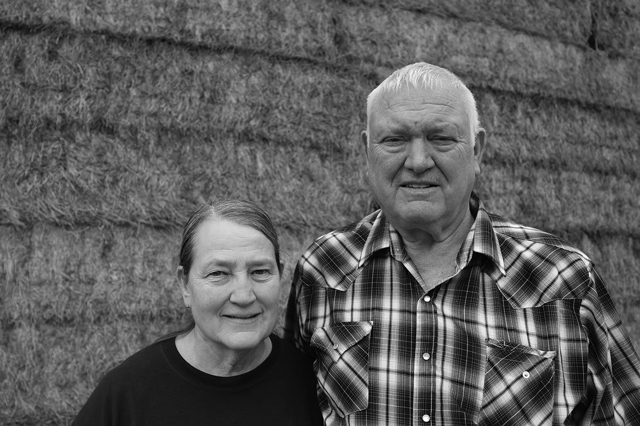 Woman in black shirt and man wearing checkered shirt standing in front of a brick building, smiling at camera