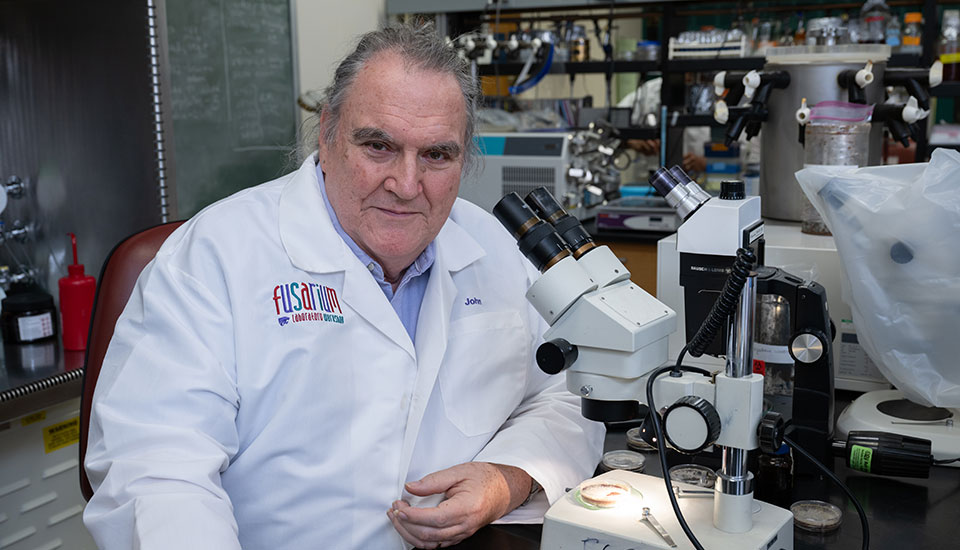 Man in white lab coat sitting in a laboratory with a microscope and other equipment in the background