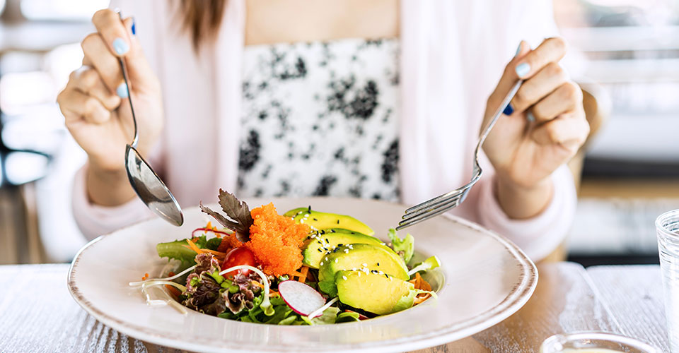 closeup of woman using two forks to eat from a white plate of various healthy foods