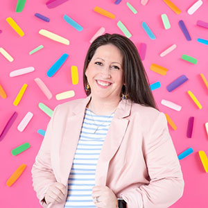 Portrait of woman with long dark hair wearing a light blazer. She is standing in front of a pink background speckled with multiple colors of candy sprinkles