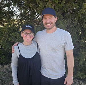 Woman and man with arms around each smiling at the camera, in front of a background of green trees