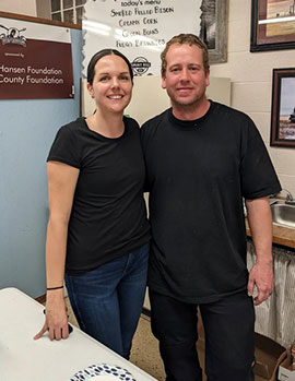 Woman and man wearing black shirts standing in a commercial kitchen, smiling at the camera