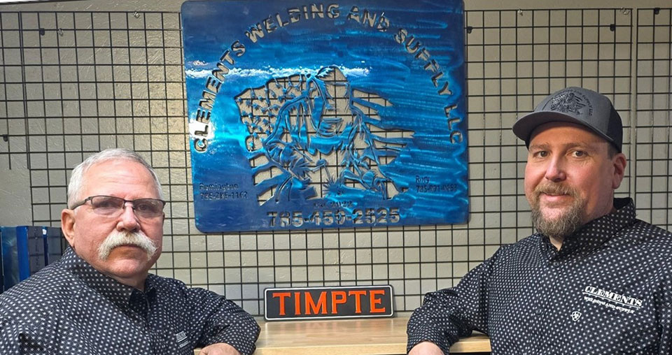 Two men in checkered shirts standing in front of counter that shows custom welded materials and signs