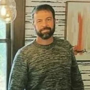 Man with beard and short hair wearing a sweater and standing against a white brick background