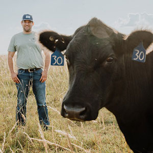 Man dressed in blue jeans and shirt standing in a farm field with a closeup of a brown cow's head in the foreground