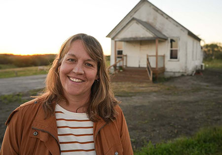 Picture of woman smiling and wearing a brown coat. The blurred background includes a white building that is a former one-room schoolhouse