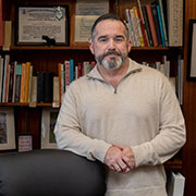 Man with a beard wearing a light brown sweater and leaning against an office chair. A shelf with several books is in the background.
